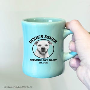 以下が含まれることがあります： ターコイズ色のダイナー風マグカップ。黒い円の中に犬の肖像画が描かれた白黒のロゴが特徴です。「Dixie's Diner」の文字が犬の上にアーチ状に配置され、その下に「Serving Love Daily」と「Est. 2013」と書かれています。