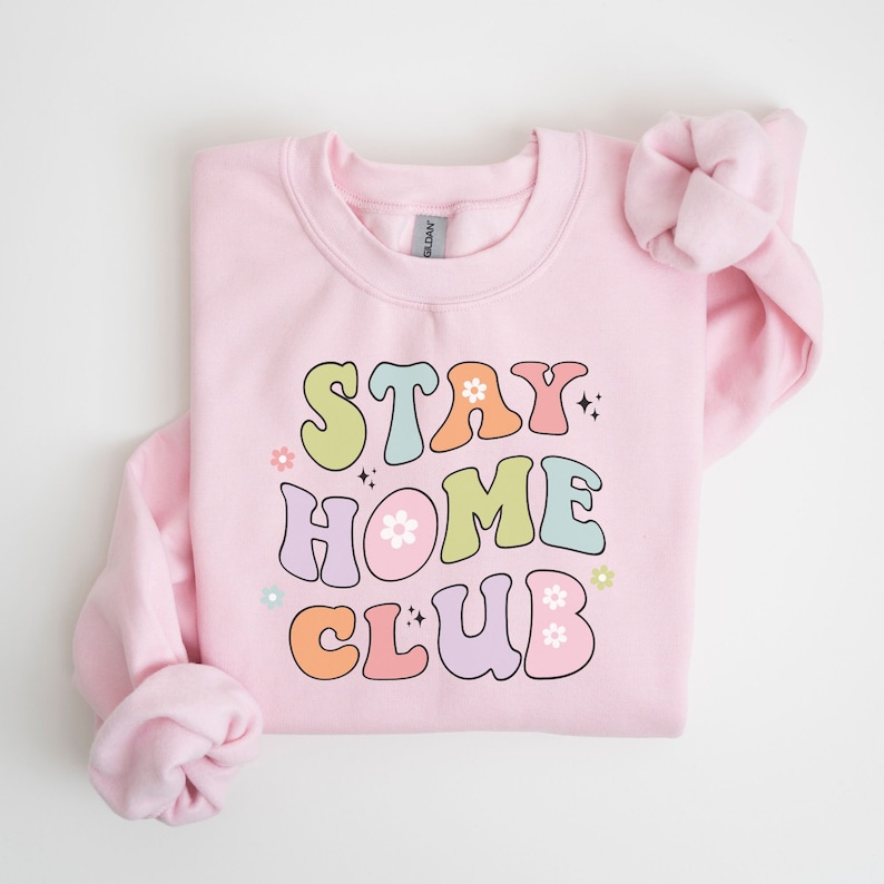 May include: Pink sweatshirt with the text "STAY HOME CLUB" in colorful retro lettering with flowers and sparkles.