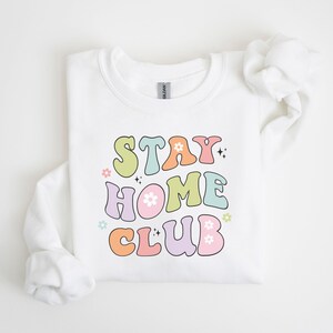 May include: White sweatshirt with a colorful retro-style graphic that says "Stay Home Club" with flowers and sparkles.
