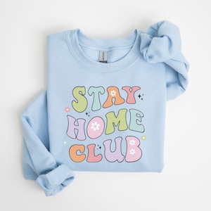 May include: Light blue sweatshirt with the text "STAY HOME CLUB" in colorful retro-style lettering with flowers and sparkles.