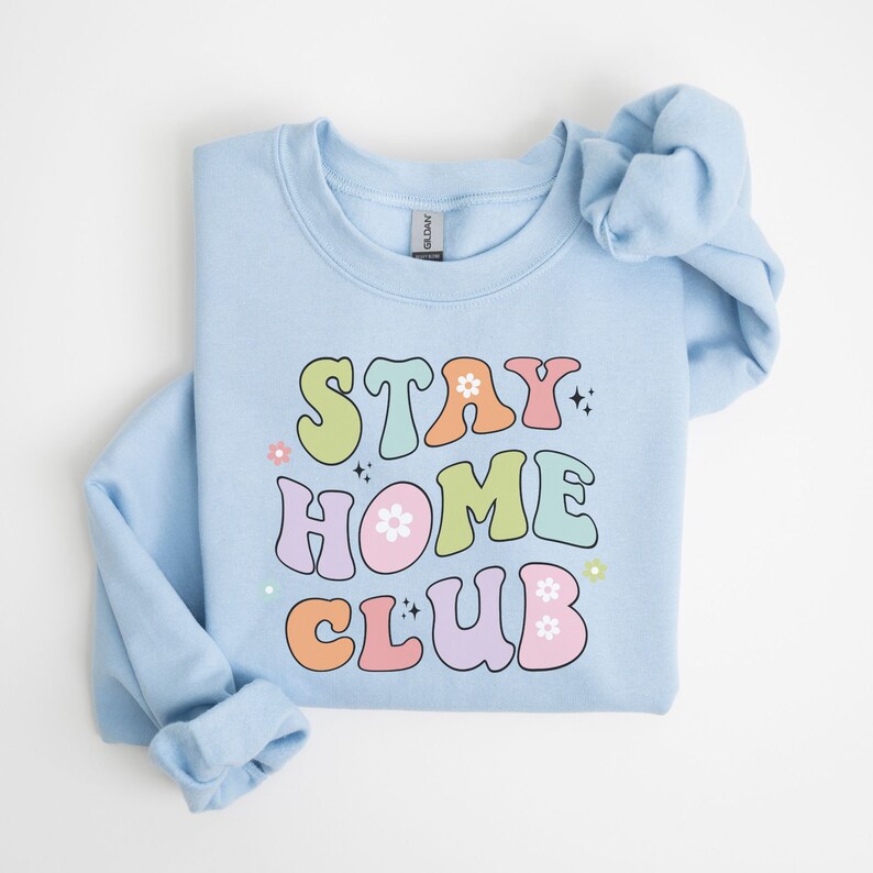 May include: Light blue sweatshirt with the text "STAY HOME CLUB" in colorful retro-style lettering with flowers and sparkles.