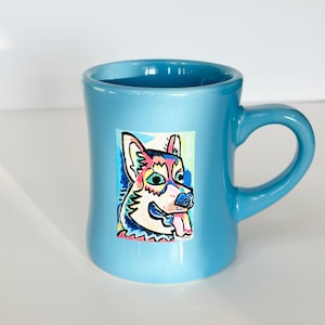 以下が含まれることがあります： 明るい青色のセラミックマグカップ。カラフルな犬のイラストが描かれています。犬はピンクの舌、青い鼻、緑の目をしています。マグカップは湾曲したハンドルと滑らかで光沢のある仕上がりです。