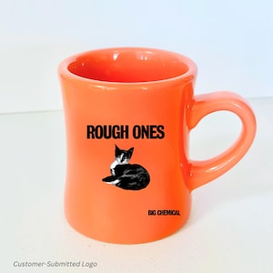 以下が含まれることがあります： オレンジ色のセラミックマグカップ、湾曲したハンドル付き。マグカップには、白黒の猫のイラストの上に「ROUGH ONES」の文字が描かれています。猫の下には「BIG CHEMICAL」の文字があります。