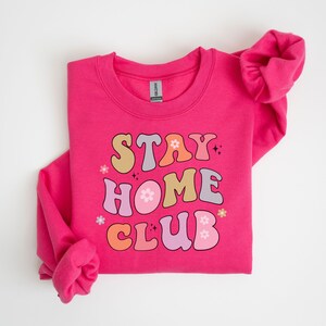 May include: Pink sweatshirt with the text "STAY HOME CLUB" in colorful retro font with flowers and sparkles.