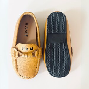 May include: A pair of tan leather loafers for children. One shoe is shown from the top, displaying the word "KULAGE" and the name "LIAM". The other shoe is shown from the bottom, with a black rubber sole. The shoes are size 25.