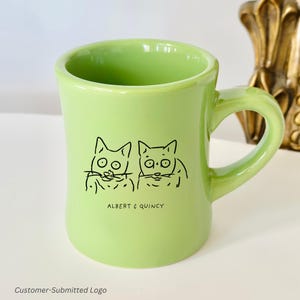 以下が含まれることがあります： 2匹の猫の黒いイラストと「ALBERT & QUINCY」の文字が入った、明るい緑色のセラミックマグカップ。マグカップはカーブしたハンドルとわずかにフレアしたリムが付いています。マグカップは明るい緑色です。