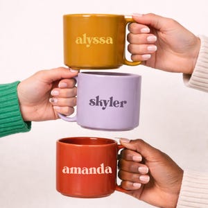 May include: Three ceramic mugs in mustard yellow, lavender, and rust red are held by different people. Each mug has a name printed on it: "alyssa", "skyler", and "amanda". The mugs have a simple, modern design with a rounded shape and a handle.