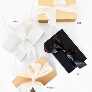 Peut inclure: Quatre bo&icirc;tes cadeaux dor&eacute;es, blanches, or rose et noires, chacune nou&eacute;e d'un ruban de satin. Les bo&icirc;tes sont dispos&eacute;es sur une surface en marbre blanc. Des &eacute;tiquettes de texte identifient chaque couleur de bo&icirc;te.