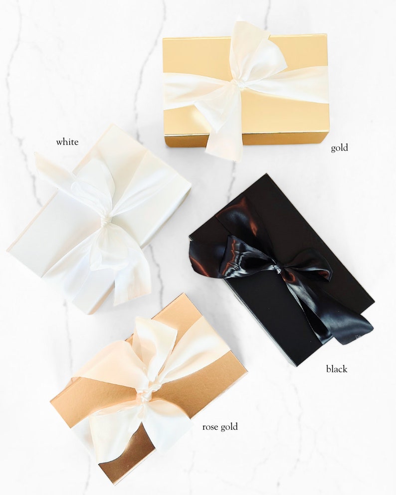 Peut inclure: Quatre bo&icirc;tes cadeaux dor&eacute;es, blanches, or rose et noires, chacune nou&eacute;e d'un ruban de satin. Les bo&icirc;tes sont dispos&eacute;es sur une surface en marbre blanc. Des &eacute;tiquettes de texte identifient chaque couleur de bo&icirc;te.