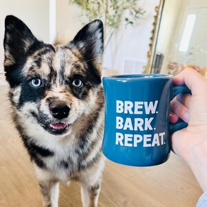 以下が含まれることがあります： 「BREW. BARK. REPEAT」の白い文字が入った青いセラミックマグが、青い目とマルチカラーの毛並みを持つ犬の近くに持たれています。犬はカメラを見ています。