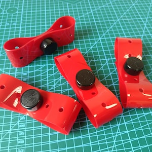 May include: Four red plastic objects with black circular components are arranged on a green grid surface. Each red piece has a curved shape with two holes and a black circular piece attached.