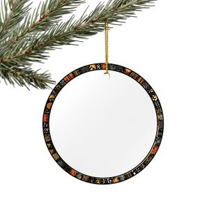 以下が含まれることがあります： オレンジ、黒、金色の模様が施された装飾的なボーダーが付いた、丸い白いクリスマスオーナメント。 オーナメントは金の紐で吊り下げられ、松の枝に飾られています。