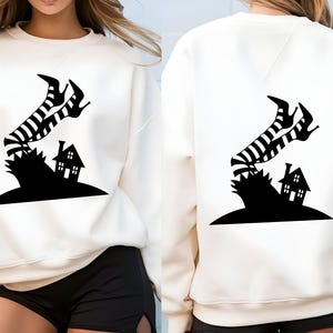 Cream-colored unisex sweatshirt featuring a black silhouette graphic of a haunted house with a witch on a broomstick flying overhead, shown on front and back views.