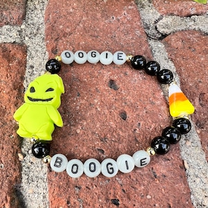 Puede incluir: Una pulsera de cuentas con las palabras "OOGIE BOOGIE" escritas con cuentas de letras blancas. La pulsera incluye cuentas negras, detalles dorados, un dije de Oogie Boogie verde y un dije de caramelo de maíz.