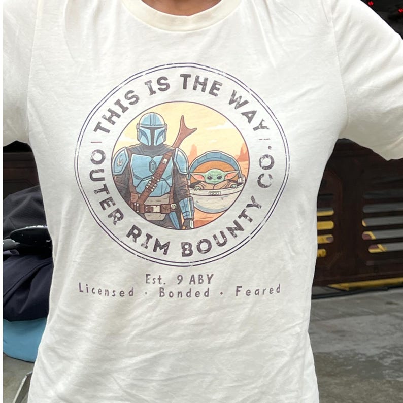 May include: White t-shirt featuring a circular graphic with the text "THIS IS THE WAY" and "OUTER RIM BOUNTY CO." The graphic includes an illustration of a character in blue armor and a small green creature. The text "Est. 9 ABY" is also present.