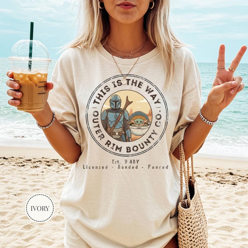 May include: Ivory-colored t-shirt featuring a circular graphic with the text "THIS IS THE WAY CO. OUTER RIM BOUNTY." The graphic includes an illustration of a Mandalorian character and Grogu. The text "Est. 9 ABY Licensed Bonded Feared" is also present.