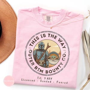 May include: A light pink t-shirt featuring a circular graphic with the text "THIS IS THE WAY CO. OUTER RIM BOUNTY." The graphic includes an illustration of a Mandalorian character and a small green creature. The shirt has a crew neck.