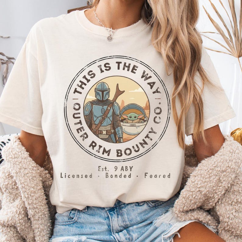 May include: Cream-colored t-shirt featuring a circular graphic with the text "THIS IS THE WAY. CO. OUTER RIM BOUNTY." The graphic includes an illustration of a character in armor and a baby-like creature. Additional text reads "Est. 9 ABY Licensed. Bonded. Feared."
