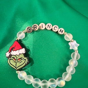 May include: A beaded bracelet featuring a Grinch charm with a red Santa hat and the word "GRINCH" spelled out in gold letter beads. The bracelet includes clear, round beads and a star-shaped charm, set against a green background.
