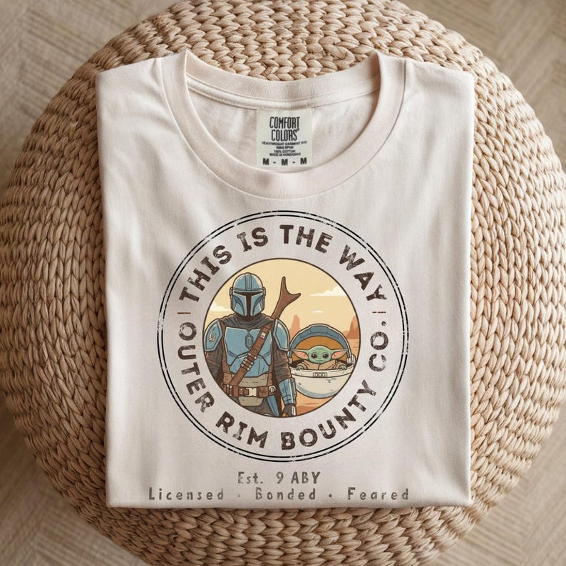 May include: Cream-colored t-shirt featuring a circular graphic with the text "THIS IS THE WAY" and "OUTER RIM BOUNTY CO." The graphic includes an illustration of a character in blue armor and a small green creature. The shirt also has the text "Est. 9 ABY".