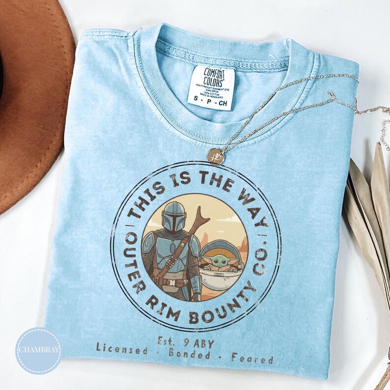 May include: Light blue t-shirt with a circular graphic featuring a Mandalorian character and Grogu, with the text "THIS IS THE WAY." The shirt also includes the text "OUTER RIM BOUNTY CO." and "Est. 9 ABY."