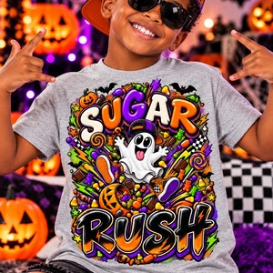 May include: A heather grey t-shirt featuring a vibrant graphic print with the text "SUGAR RUSH" in bold, colourful lettering. The design includes a cartoon ghost wearing a cap, surrounded by various Halloween sweets, pumpkins, and bats.