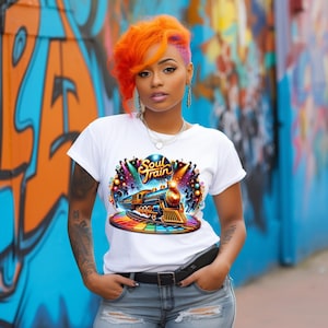 May include: White t-shirt featuring a colourful graphic of a train, the words "Soul Train", and a rainbow-coloured platform. The image is set against a graffiti-covered wall. The person is wearing blue jeans and a black belt.