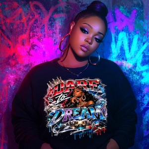 May include: Black sweatshirt with the words "DARE to DREAM" in a stylized design. The graphic includes an illustration of a woman, a crown, and city skyline elements. The background features graffiti art in shades of pink, blue, and purple.