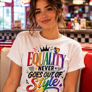 Puede incluir: Una camiseta blanca de manga corta con un estampado gráfico colorido que dice "EQUALITY NEVER GOES OUT OF STYLE" en letras de colores del arcoíris, acompañada de una corona, un corazón y un icono de percha con un corazón arcoíris.