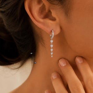 May include: A silver-toned hoop earring featuring a vertical drop design with five small, clear, round-cut stones arranged in a descending size order, suspended from a small hinged hoop that secures through the earlobe.
