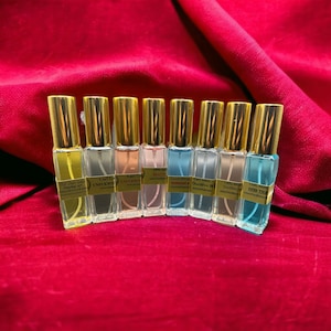 May include: A collection of eight perfume bottles with gold caps and clear glass bodies, each containing a different colored liquid. The bottles are arranged on a red velvet surface. Each bottle has a label with text.