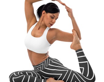 May include: A person wearing a white sports bra and black leggings with a white geometric pattern, in a yoga pose. The leggings have a zig-zag design. The background is white.