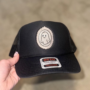 May include: A black trucker hat featuring a central embroidered patch of a white ghost inside an ornate oval frame. The hat includes a black foam front panel, a mesh back, and a sticker on the brim that reads OTTO WORLD FAMOUS FOAM FRONT OSFM TRUCKER.
