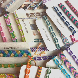 May include: A collection of handcrafted friendship bracelets in various colors and patterns. The bracelets are displayed on small cardboard tags, some with the word "HANDCRAFTED" printed on them. The bracelets feature woven designs in shades of pink, green, blue, orange, and purple.