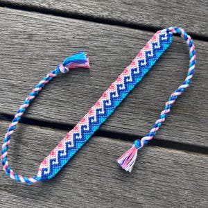 Puede incluir: Una pulsera de amistad hecha a mano con un patrón geométrico azul, rosa y blanco. La pulsera tiene cordones trenzados y borlas en cada extremo. La pulsera está sobre una superficie de madera.