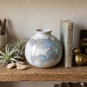 May include: A round, off-white ceramic vase with a blue and purple iridescent glaze. The vase sits on a wooden shelf with a small book, a brass bird figurine, and air plants. The book has the words "Handwriting Notebook" on the spine.