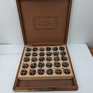 May include: A wooden storage box containing a set of metal letter stamps for a Kingsley Gold Stamping Machine. The interior lid features the text KINGSLEY Gold Stamping Machine Co. Hollywood California. The base has circular compartments.