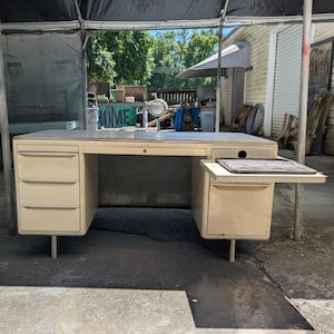 May include: A beige metal desk featuring a flat rectangular top, three stacked drawers on the left side, and a single drawer on the right side. A pull-out shelf with a patterned surface is extended from the right side above the bottom drawer.