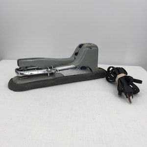 May include: A gray electric stapler with a black base and a silver metal staple guide. The stapler has a long, curved handle and a black power cord with a tan rubber band wrapped around it.