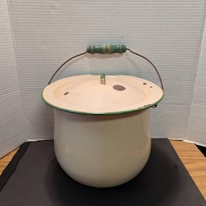 May include: Cream-colored enamelware pot with a lid and a wire handle. The pot has a green rim and a green wooden handle. The lid has a small handle and some dark spots. The pot is likely vintage or antique.