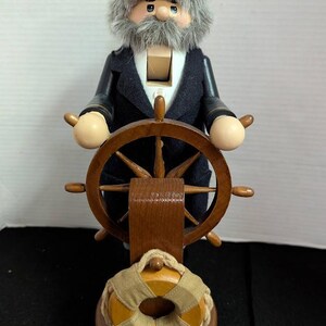 May include: A wooden nutcracker in the shape of a sea captain, wearing a navy blue jacket and a tan cap with an anchor emblem. The captain figure has a gray beard and holds a wooden ship's wheel. A life preserver is at the base.