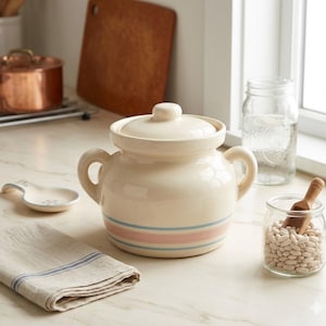 May include: Cream-colored ceramic bean pot with a lid, two handles, and blue and pink stripes. A small glass jar with white beans and a wooden scoop is next to the pot. A folded linen towel and a spoon rest are also visible.