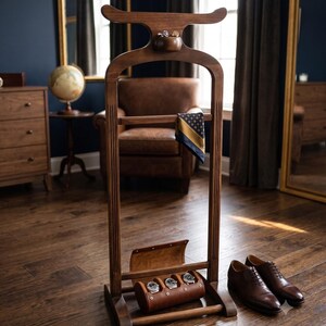 May include: A dark brown wooden valet stand featuring a curved top hanger, a small integrated bowl for accessories, a horizontal bar draped with a patterned pocket square, and a lower leather storage case holding three wristwatches.