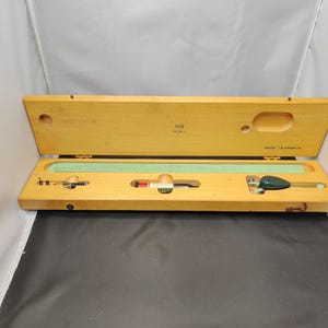 May include: A vintage drafting set in an open wooden case. The case is light brown with a hinged lid and contains a ruler, compass, and other tools. The text "K+E DORIC" is visible on the lid. The set is likely used for technical drawing or engineering.