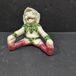 May include: A ceramic figurine of a seated elf with a smiling face. The elf wears a white outfit with green leaf accents and red shoes. The figurine has a whimsical design and is set against a dark background.