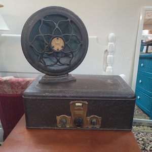 May include: An antique radio with a dark brown, textured cabinet and a large, circular speaker. The speaker has a decorative, dark metal grill and a central emblem. Brass knobs and controls are visible on the front of the radio.