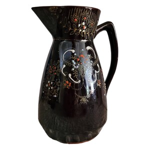 May include: A dark brown ceramic pitcher featuring a glossy finish and a decorative hand-painted floral pattern in white, orange, and gold. The pitcher has a curved handle, a wide base, and a prominent pouring spout, with intricate line work.