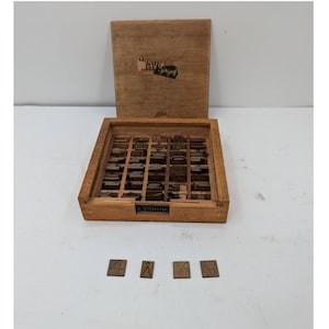 May include: A wooden storage box with a hinged lid featuring the text Gravo Graph, containing a grid of small metal letter and number stamps. A metal plate on the front reads A 1200/15. Four individual metal stamps showing 4, A, 7, and 9 lie in front.