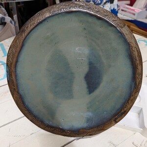 May include: A handmade ceramic plate with a wide, decorative rim. The plate's interior has a mottled blue-green glaze, with darker blue areas. The rim is a contrasting brown with an embossed floral pattern. The plate is on a white wooden surface.