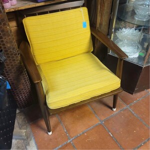 May include: A mid-century modern armchair with a bright yellow cushion and backrest. The chair has a wooden frame with dark brown armrests and legs. The legs have white tips. The chair is placed on a terracotta tile floor.